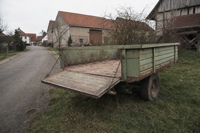 Leerer zweirädriger Anhänger mit heruntergeklappter Klappe auf einem Grünstreifen an einer Dorfstraße