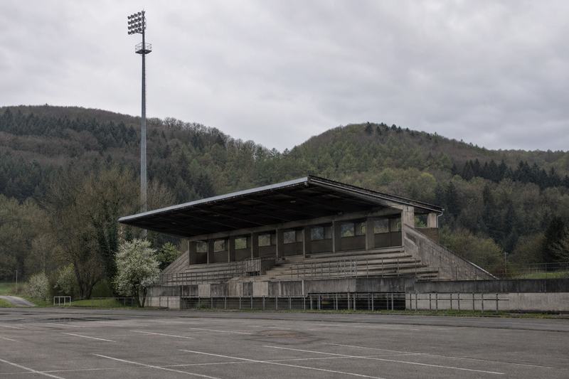 Betontribüne eines Fußballplatzes von außen, Flutlichtmast und bewaldete Hügel dahinter