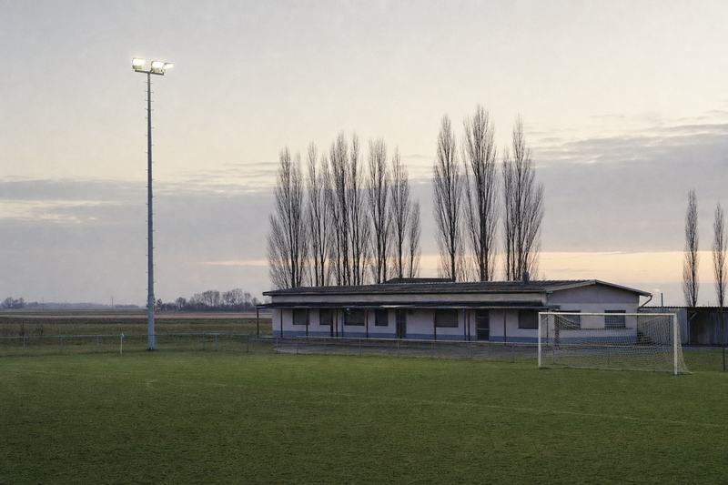 Leerer Dorfsportplatz in der Abenddämmerung, ein beleuchteter Flutlichtmast und das Vereinsheim dahinter