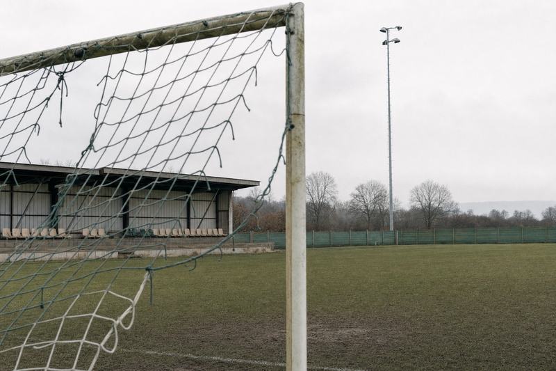 Blick hinter das Tor eines Amateurplatzes mit niedriger Tribüne und Flutlichtmast