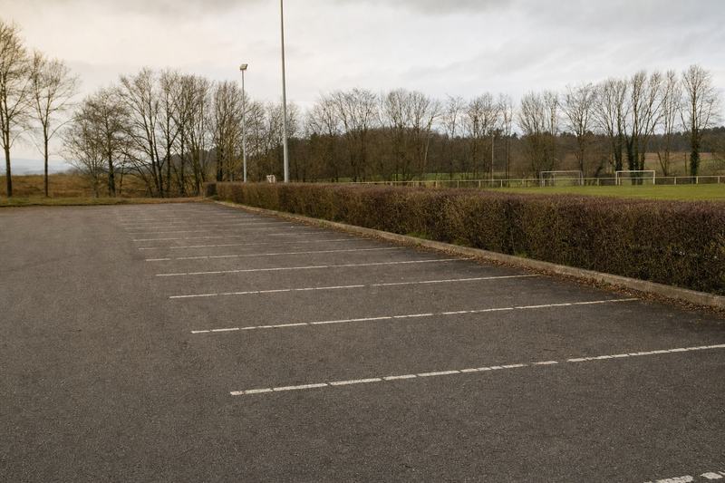 Leerer asphaltierter Parkplatz neben einem Sportgelände, ausgeblichene Markierungen