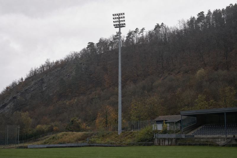 Einzelner Flutlichtmast vor einem steilen bewaldeten Hang über einem kleinen Stadion