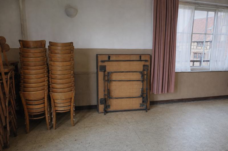 Gestapelte Holzstühle an einer verputzten Wand in einem Dorfsaal, daneben ein zusammengeklappter Klapptisch